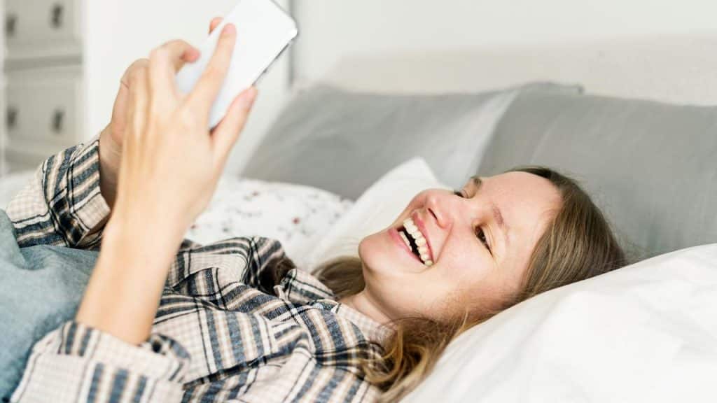 A woman smiling while lying in bed and looking at her phone.