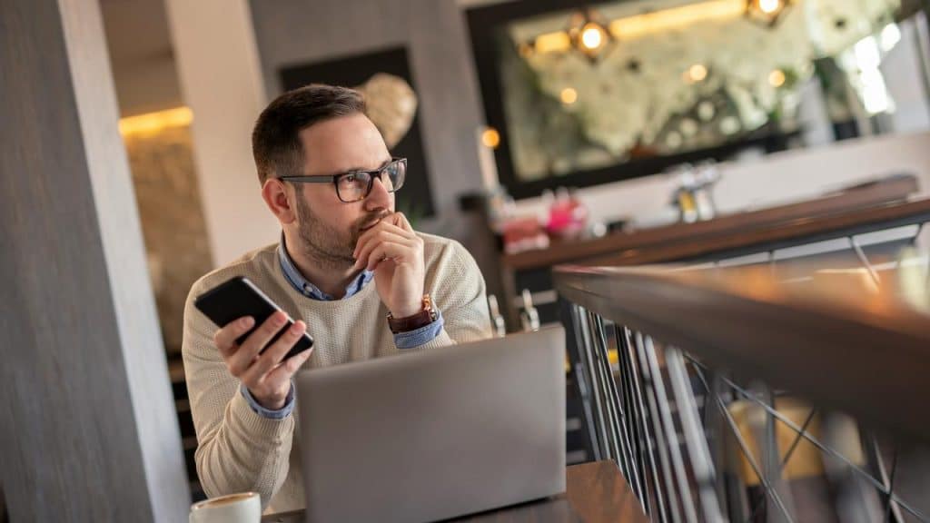 A man sitting at a table with a laptop holding a phone and looking thoughtful.