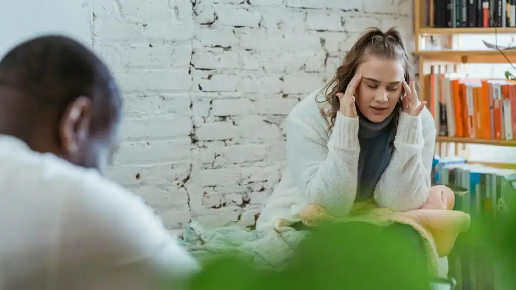 A woman sits with her hands on her temples while a man sits blurred in the foreground.