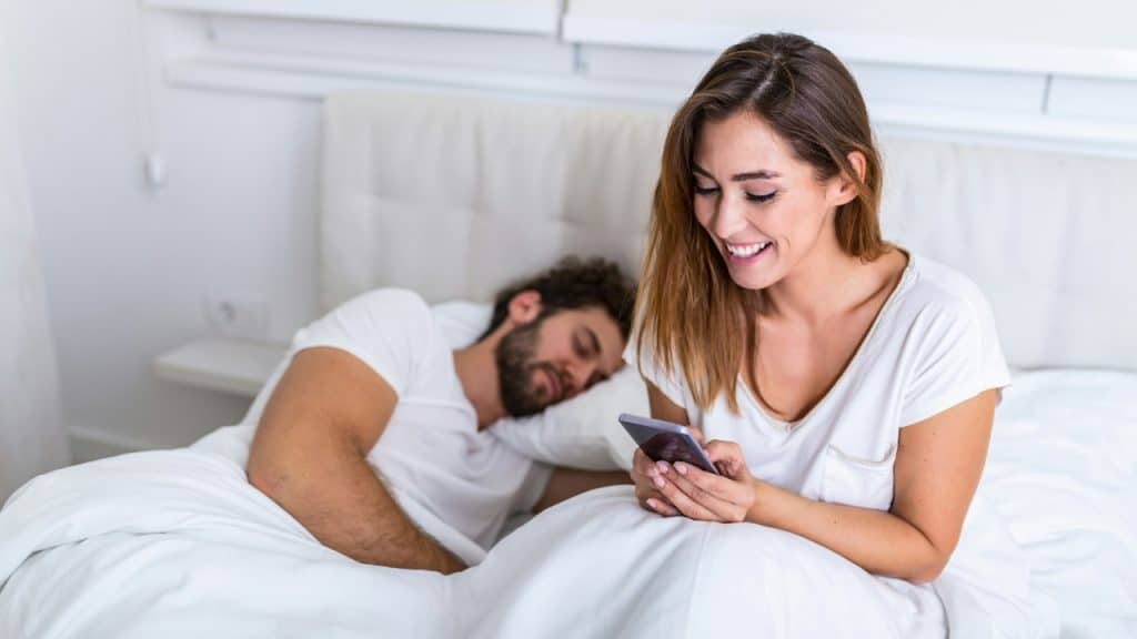 A smiling woman looks at a phone in bed while a man sleeps in the background.