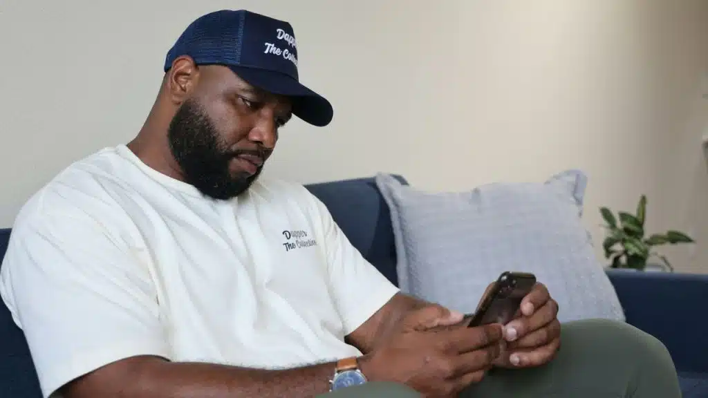 A bearded man wearing a baseball cap sits on a couch while looking at a smartphone.
