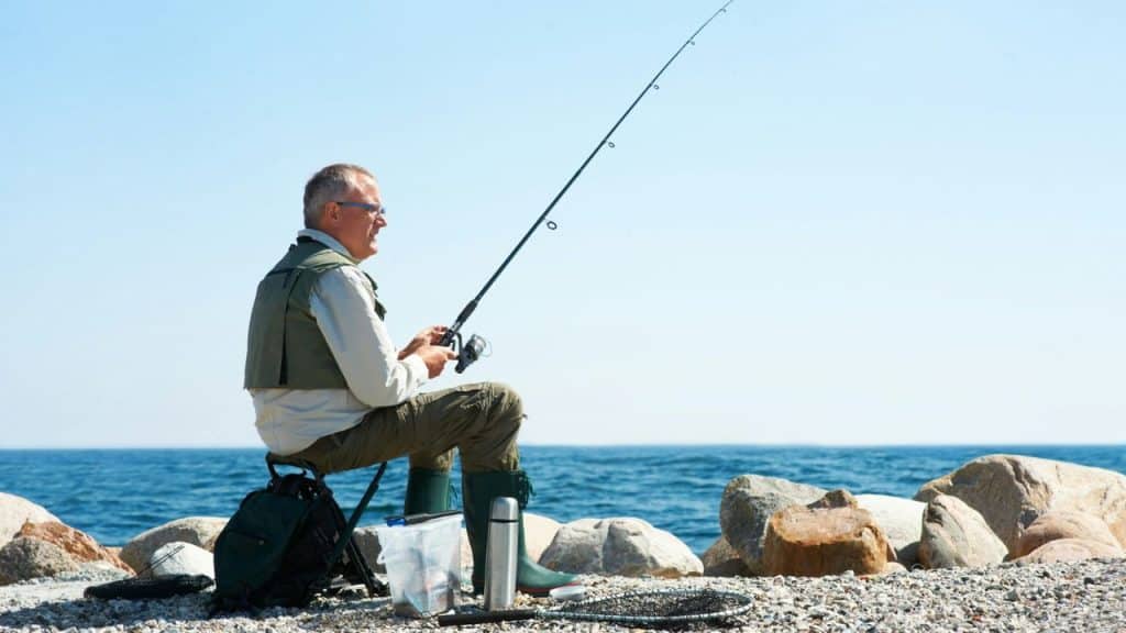 A man sitting on a rocky shore while holding a fishing rod over the water.