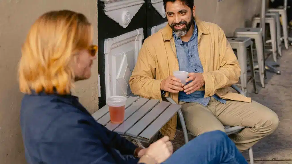 A man smiles while talking with a friend at an outdoor table.