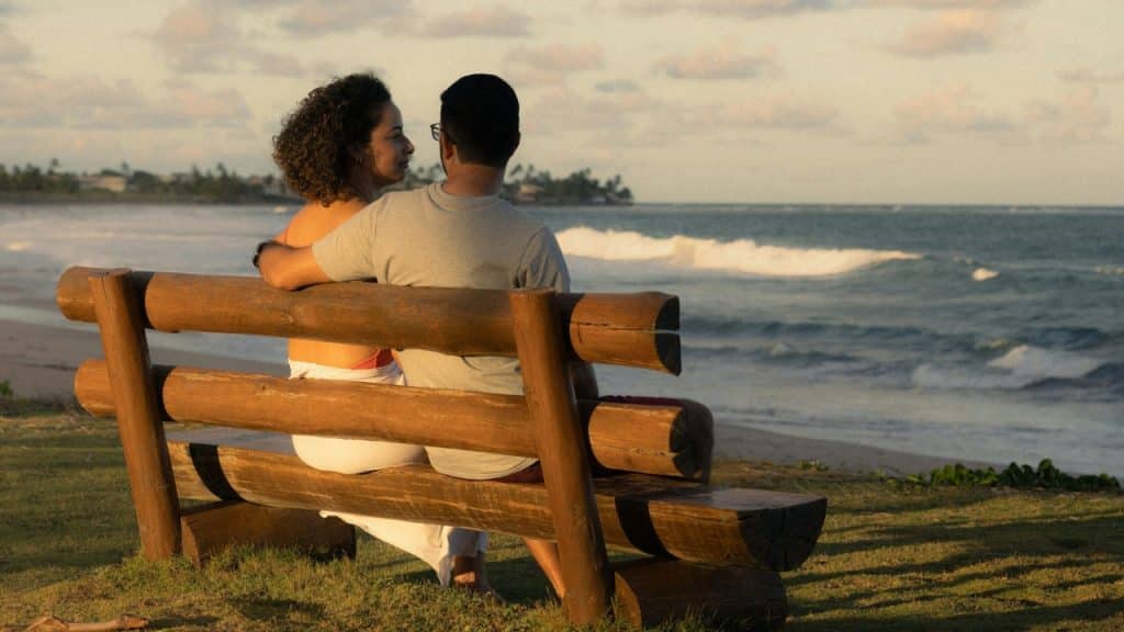 A man and woman sit on a wooden bench facing the ocean during a sunset.