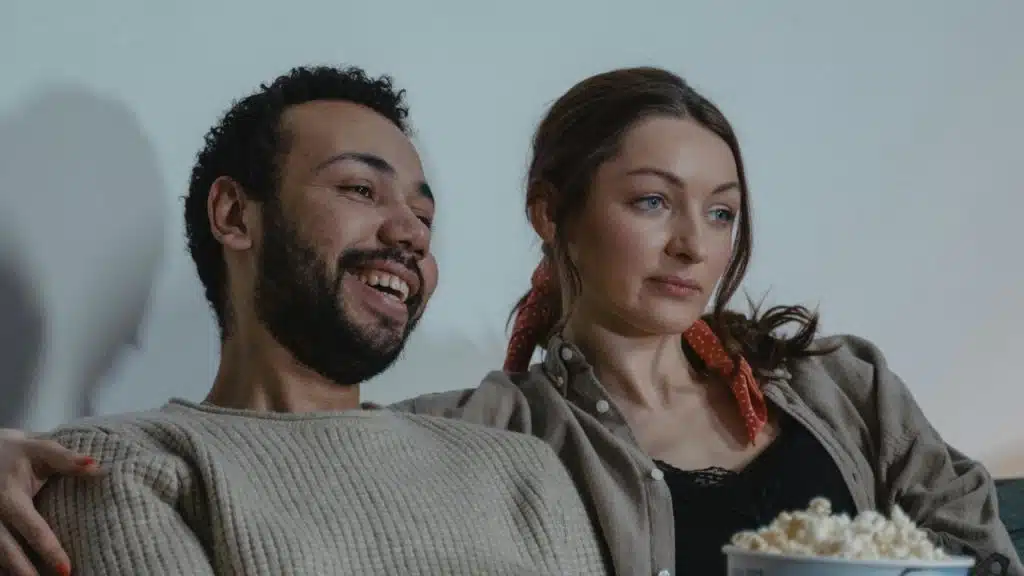 A man and woman sit together on a couch; the man laughs while holding popcorn.