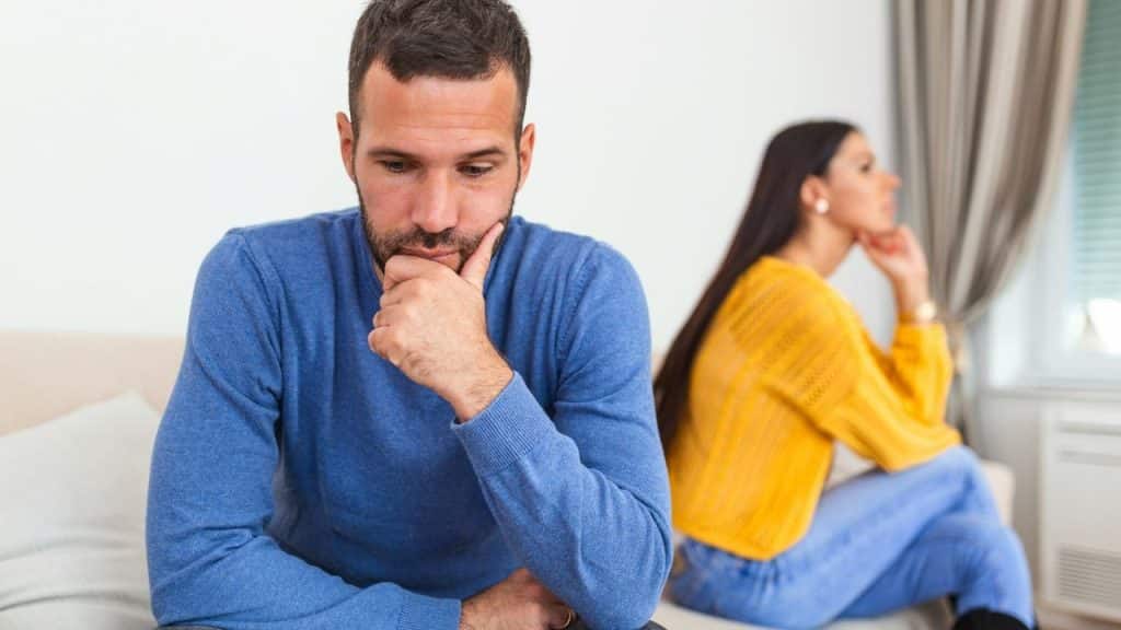 A man in a blue sweater looks down while a woman sits behind him facing away.