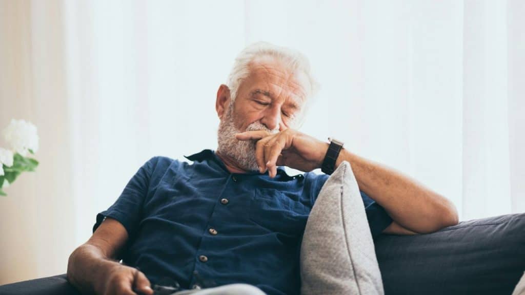 An older man with white hair and a beard sits on a couch resting.