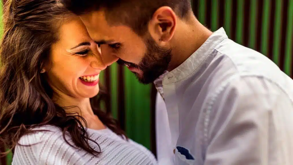 A smiling man and woman lean their foreheads together in front of a green background.
