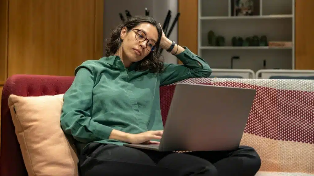 A woman wearing glasses and a green shirt looks at a laptop on a couch.