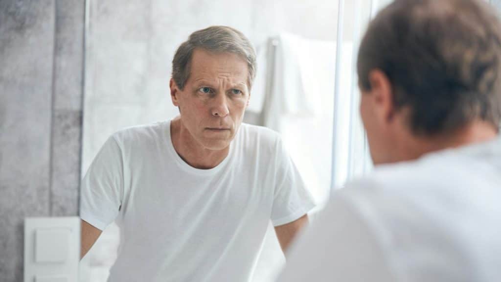 A middle-aged man looks at his reflection in a bathroom mirror with a serious expression.
