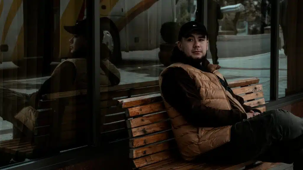 A man wearing a vest and cap sits on a wooden bench outdoors near glass.