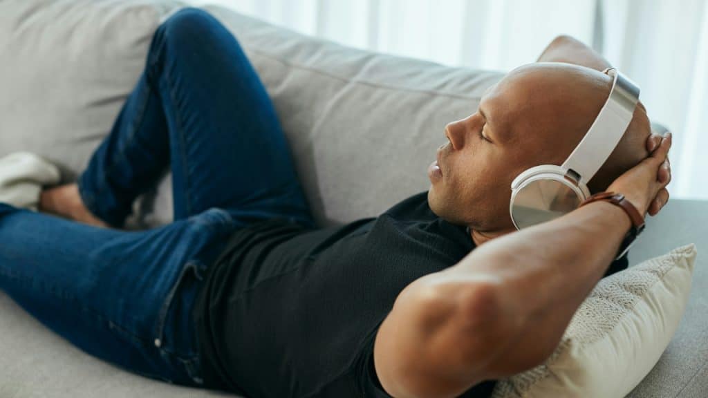 A man lies on a grey sofa wearing white headphones with his eyes closed comfortably.