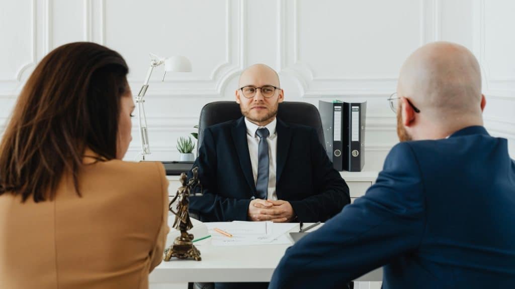 A man in a suit sits behind a desk facing two people in an office.