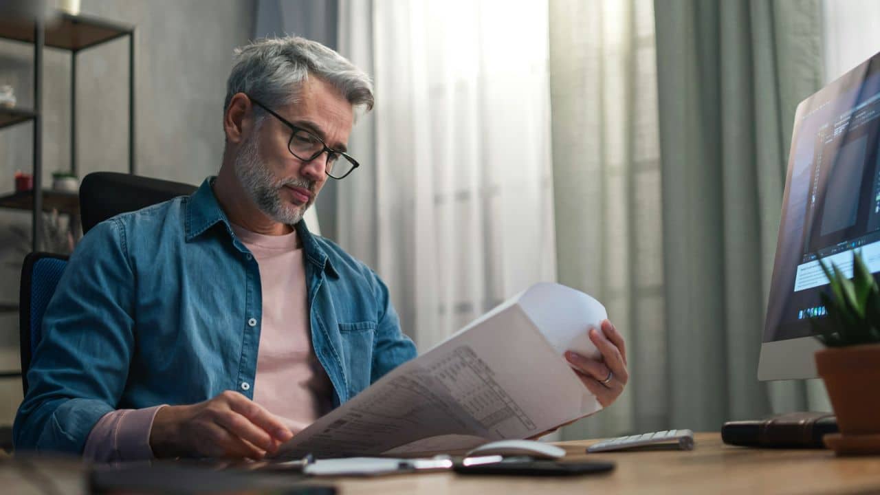 A man with gray hair and glasses reviews paper documents at a desk with a computer.