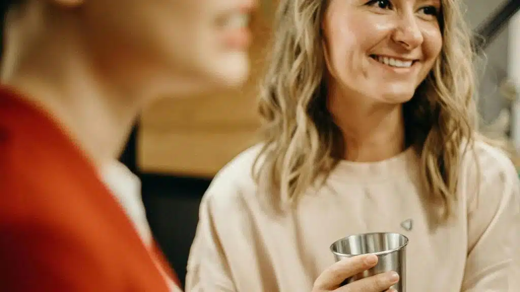 A smiling woman holding a metal cup while chatting with a friend.