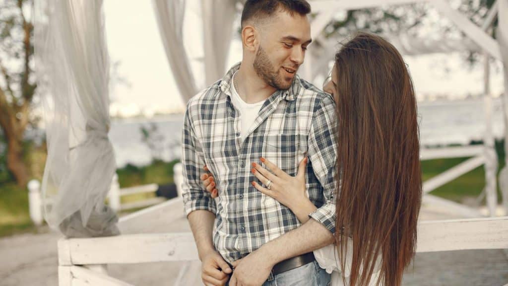 A couple smiling and embracing outdoors near a waterfront.