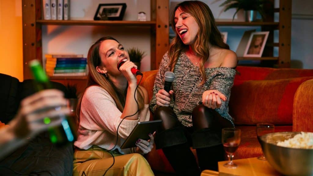 A pair of women singing karaoke together on a couch at home.