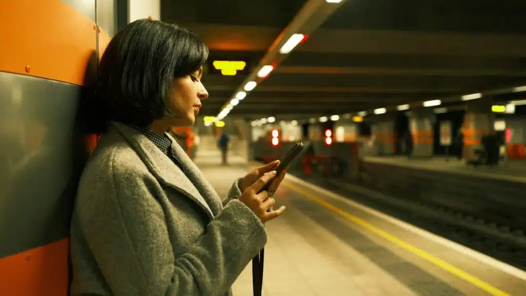 A woman in a grey coat stands in a subway station looking at her smartphone.