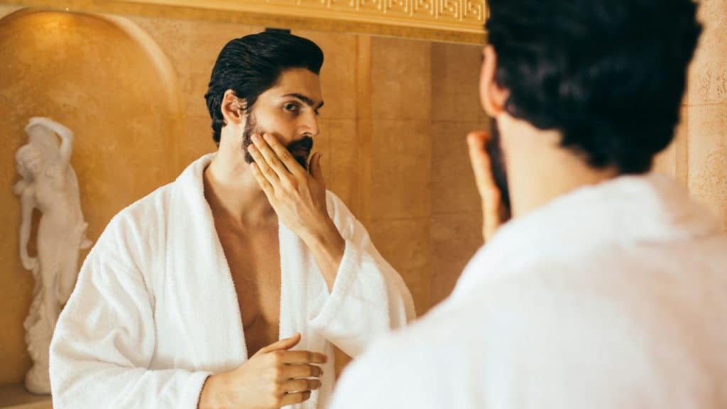 A man in a white robe looks into a mirror and touches his bearded face.