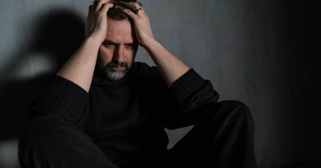 A distressed man sitting with his hands on his head in a dark room.