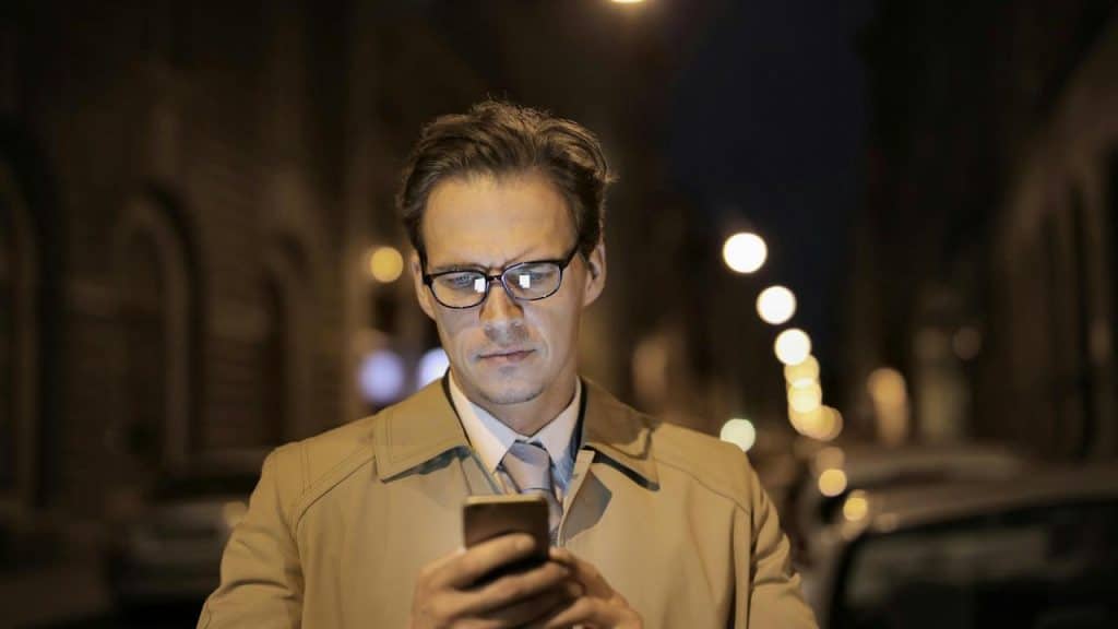 A man standing on a dimly lit street at night while looking at his phone.
