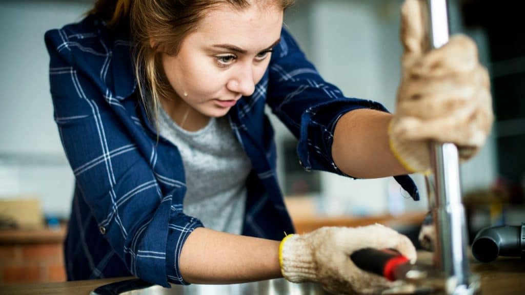 A woman wearing work gloves uses a wrench to tighten a metal pipe in a kitchen.