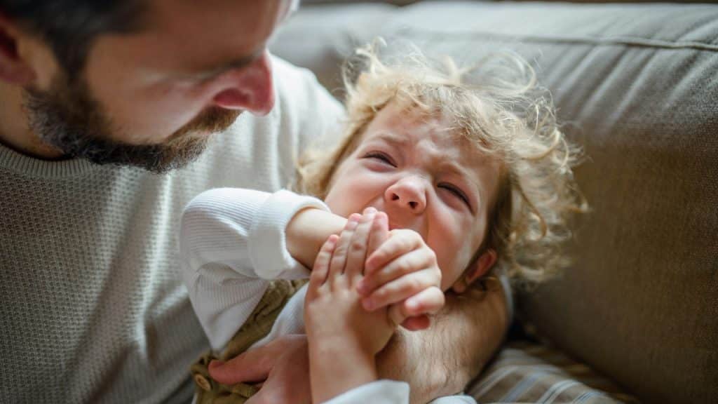 A man with a beard holds a young child who is crying with their eyes closed.