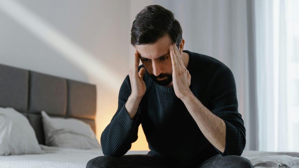 A bearded man in a dark sweater sits on a bed holding his temples with both hands.