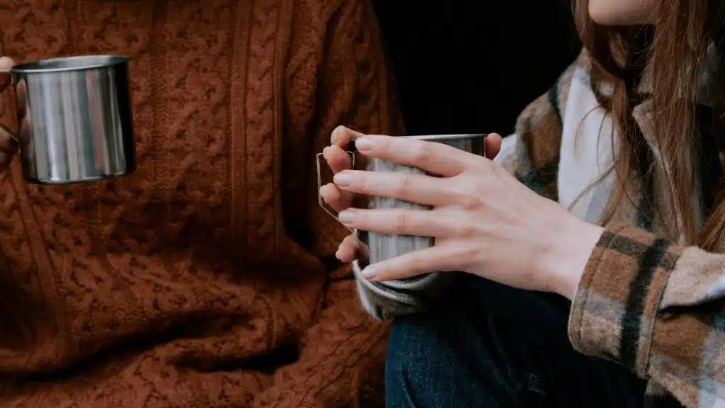 A close-up of two people holding metal mugs while wearing cozy sweaters.