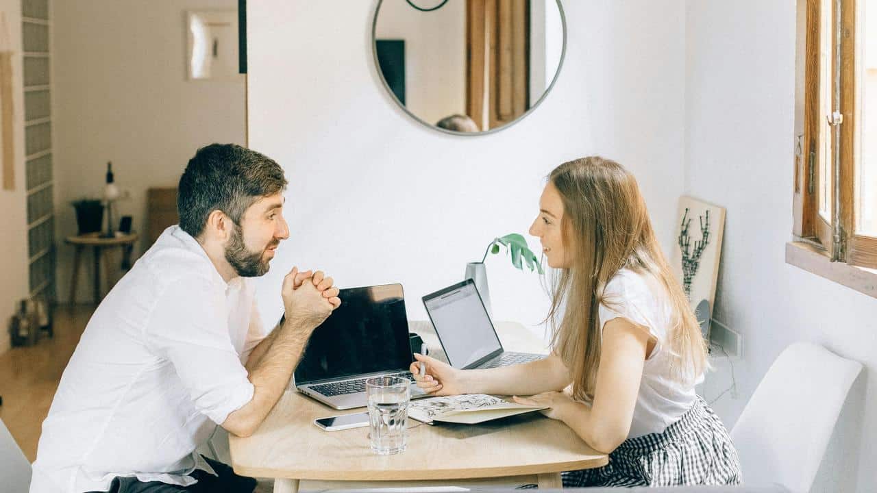 A man and woman smiling while talking and working on laptops at a table.
