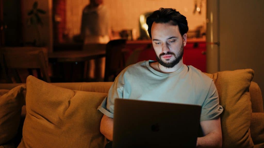 A bearded man sits on a couch at night using a glowing laptop in a room.