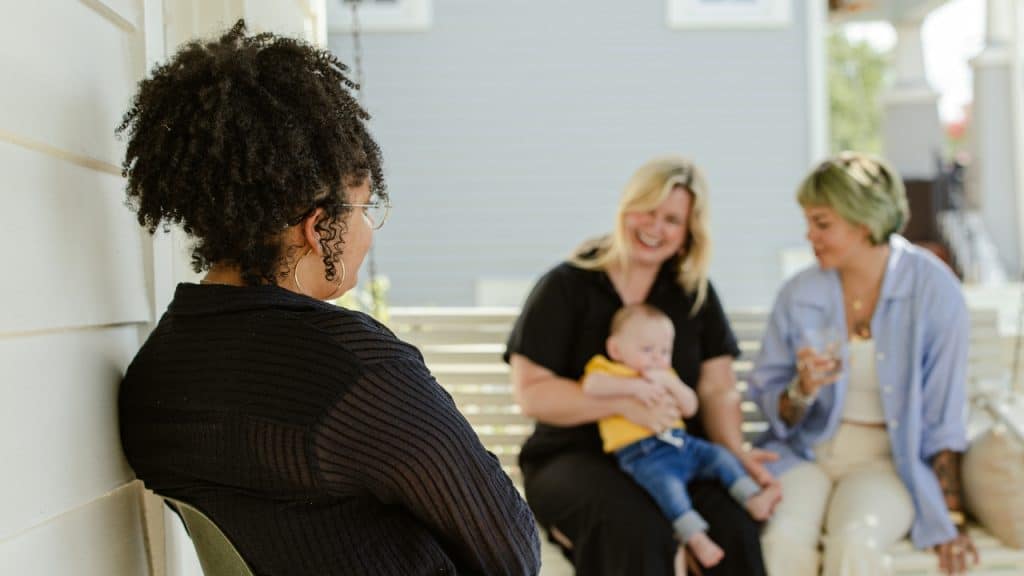 A woman in a dark shirt looks toward two people sitting on a porch swing.