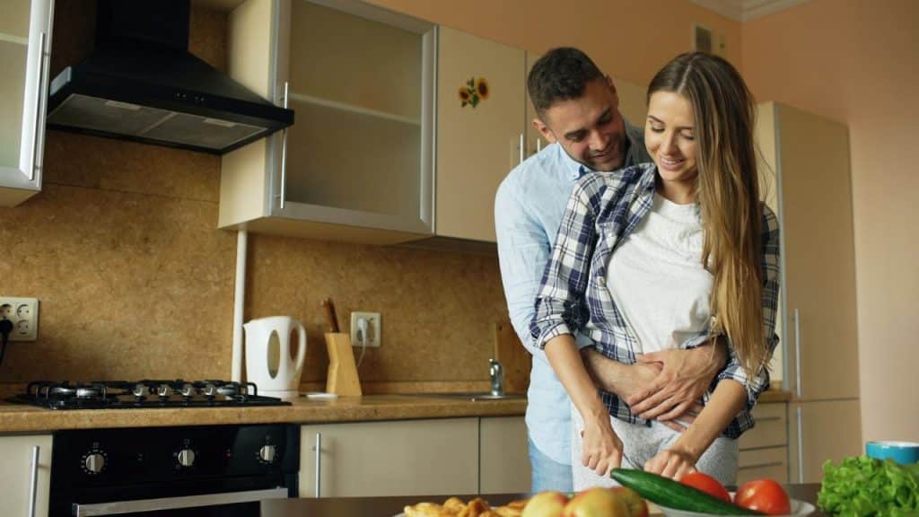 A man hugs a woman from behind as she prepares food at a kitchen counter.