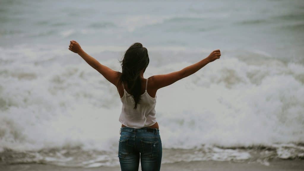 Rear view of a woman standing with arms outstretched facing the crashing ocean waves.