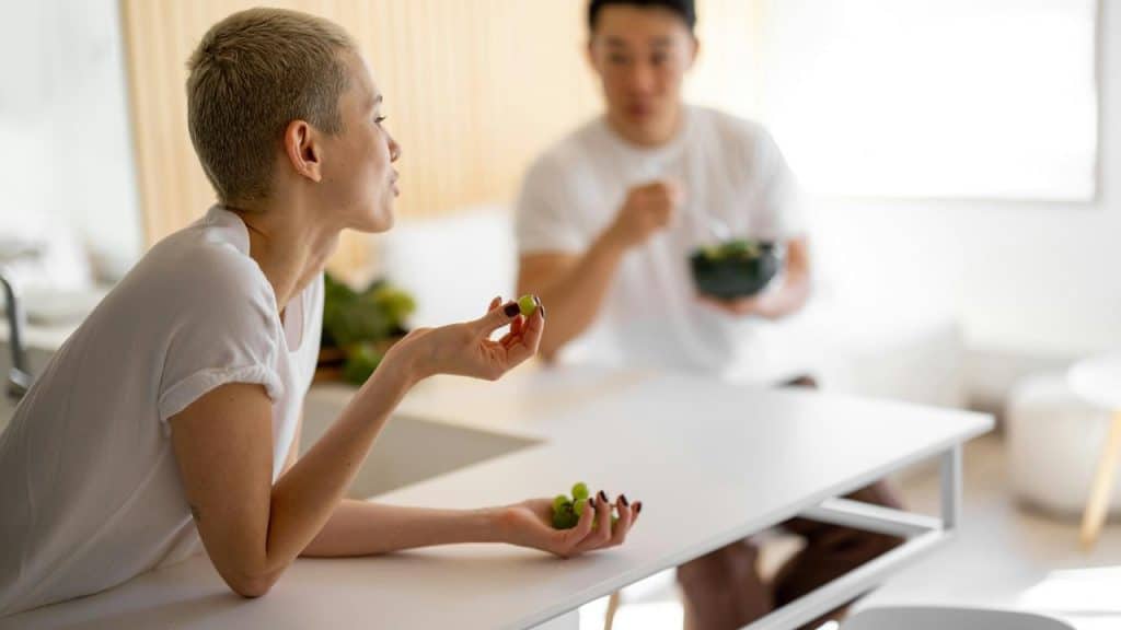 A person eating grapes while talking with another person at a table indoors.