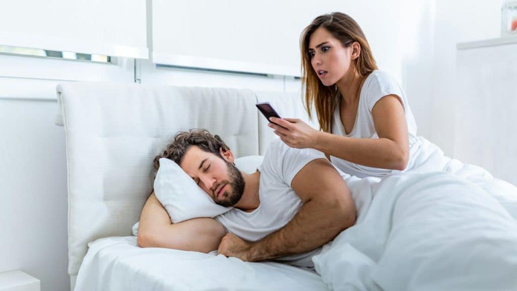A woman with an angry expression looks at a phone while a man sleeps in bed.