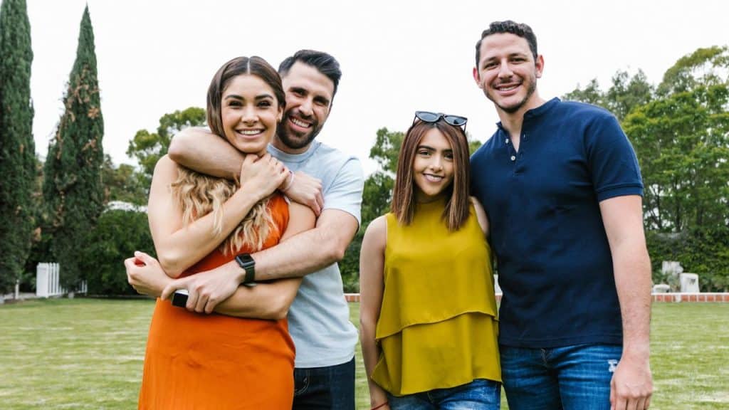 Two couples stand and smile together on a grassy lawn with trees in the background.