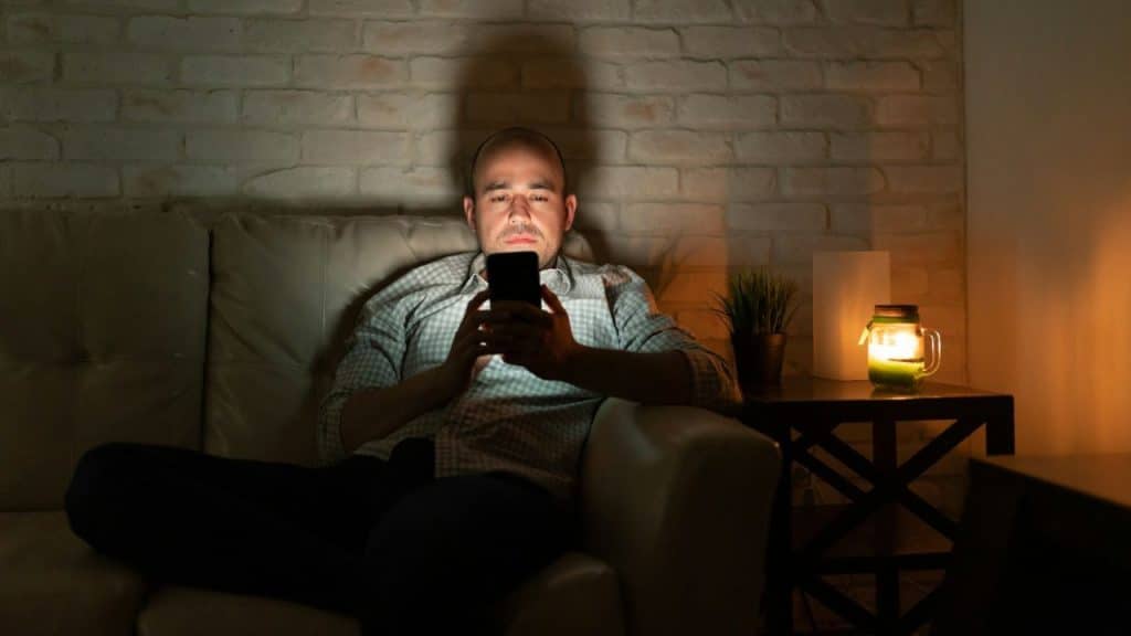 A man sits on a couch in a dark room looking at his glowing smartphone screen.