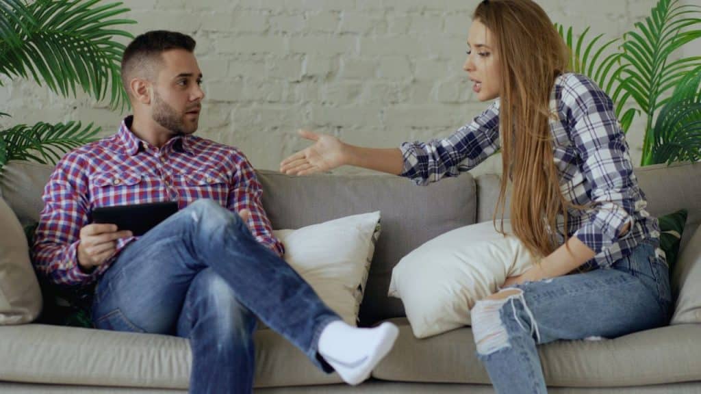 A woman gestures with her hand while talking to a man sitting on a sofa with a tablet.