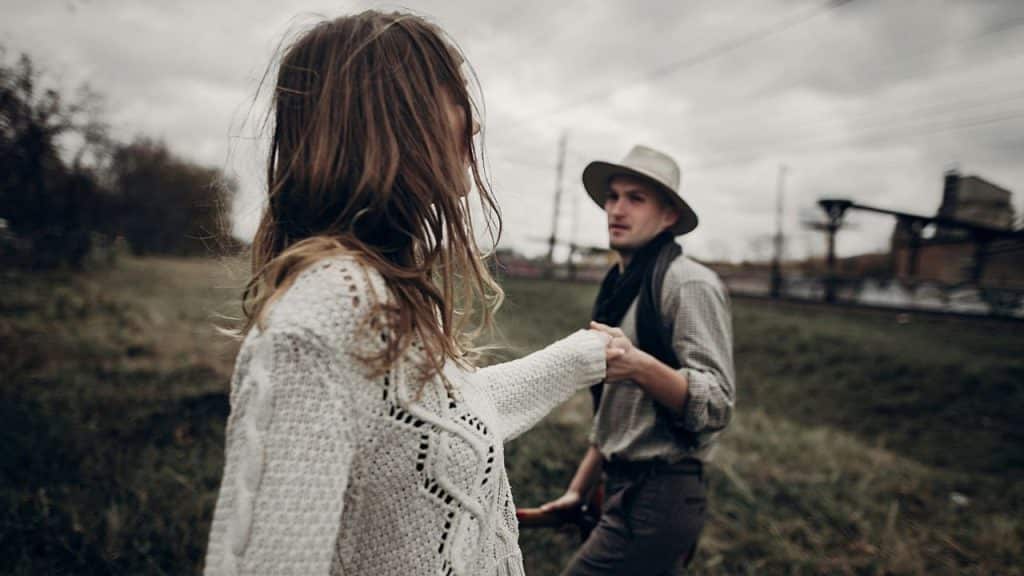 A woman in a white sweater holds hands with a man in a hat outdoors.