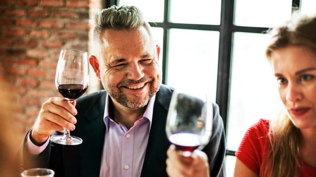 A smiling man in a suit jacket holds up a glass of red wine indoors.