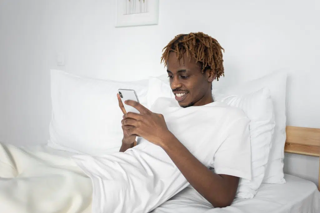 A Man in White Shirt Using a Smartphone while Lying on the Bed