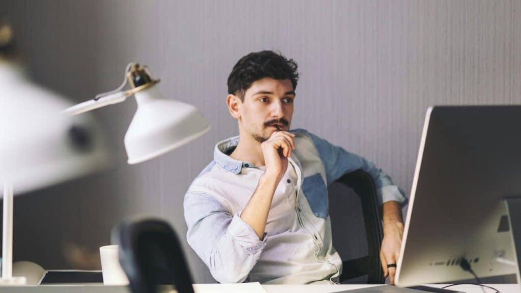 A man sitting at a desk, thoughtfully looking at a computer screen.
