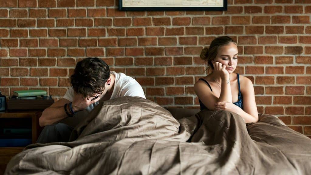 A man and woman sit in a bed against a brick wall looking in opposite directions.