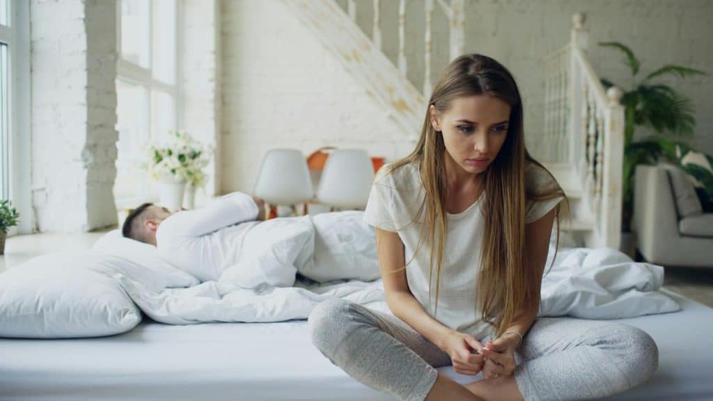 A woman sits on a bed looking down while another person lies in the background.