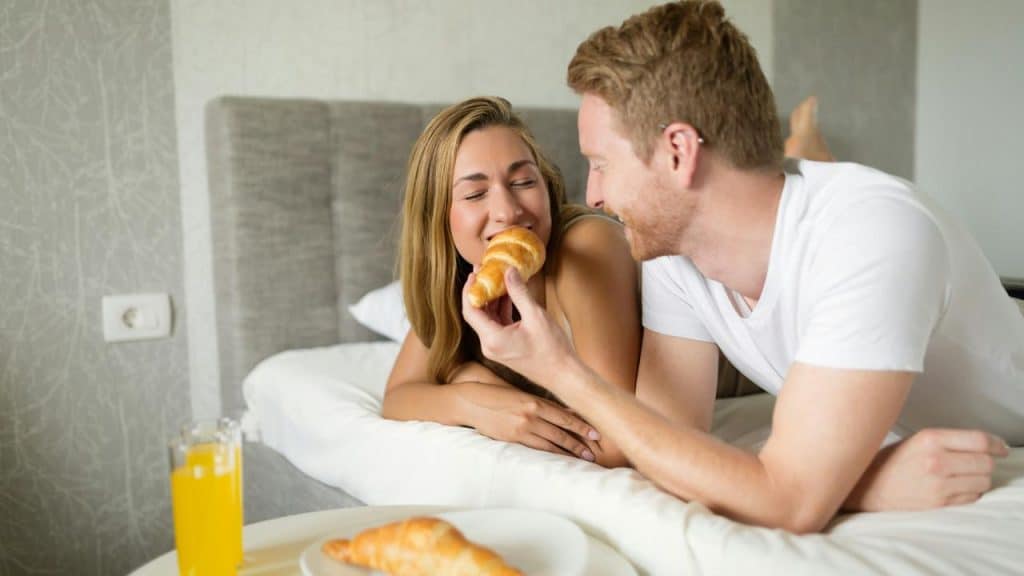 A couple in bed smiling as one offers a croissant for breakfast.