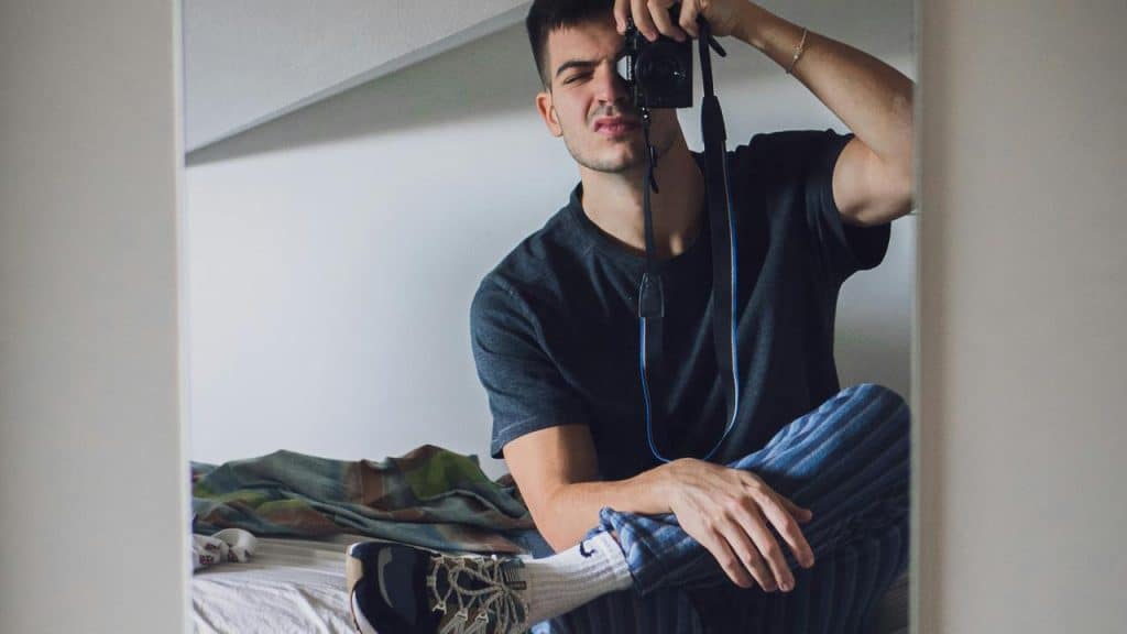 A man sits on a bed taking a mirror selfie with a camera.