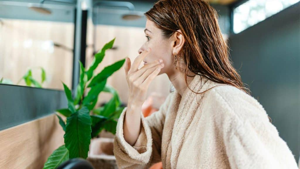 A woman in a bathrobe applying skincare while looking in the mirror.