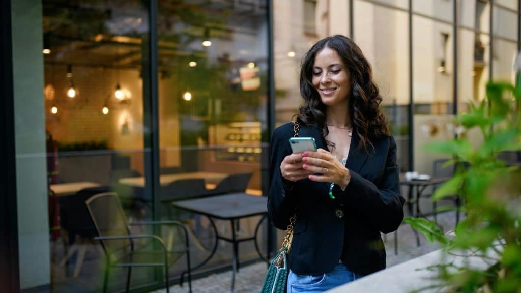 A woman in a black blazer smiles while looking at her phone outdoors.