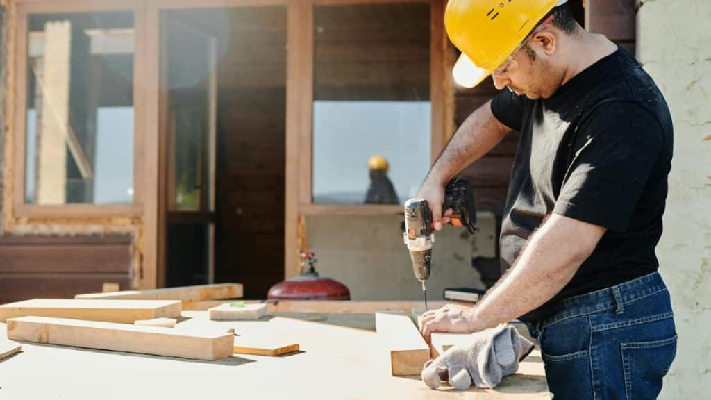 A man in a yellow hard hat uses a power drill on wood planks outdoors.